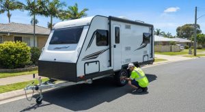 modern caravan parked on a residential Gold Coast street or driveway, with a licensed mobile roadworthy inspector in hi-vis uniform examining the wheels, brakes, or tow hitch. Include bright, natural daylight with a relaxed suburban coastal backdrop — think palm trees, clear skies, and a quiet neighbourhood. The caravan should appear well-maintained, with the focus on professionalism and safety. The scene should evoke trust, mobility, and convenience, reflecting a local, friendly roadworthy service in action. No text or logos — just an authentic, visually engaging moment.